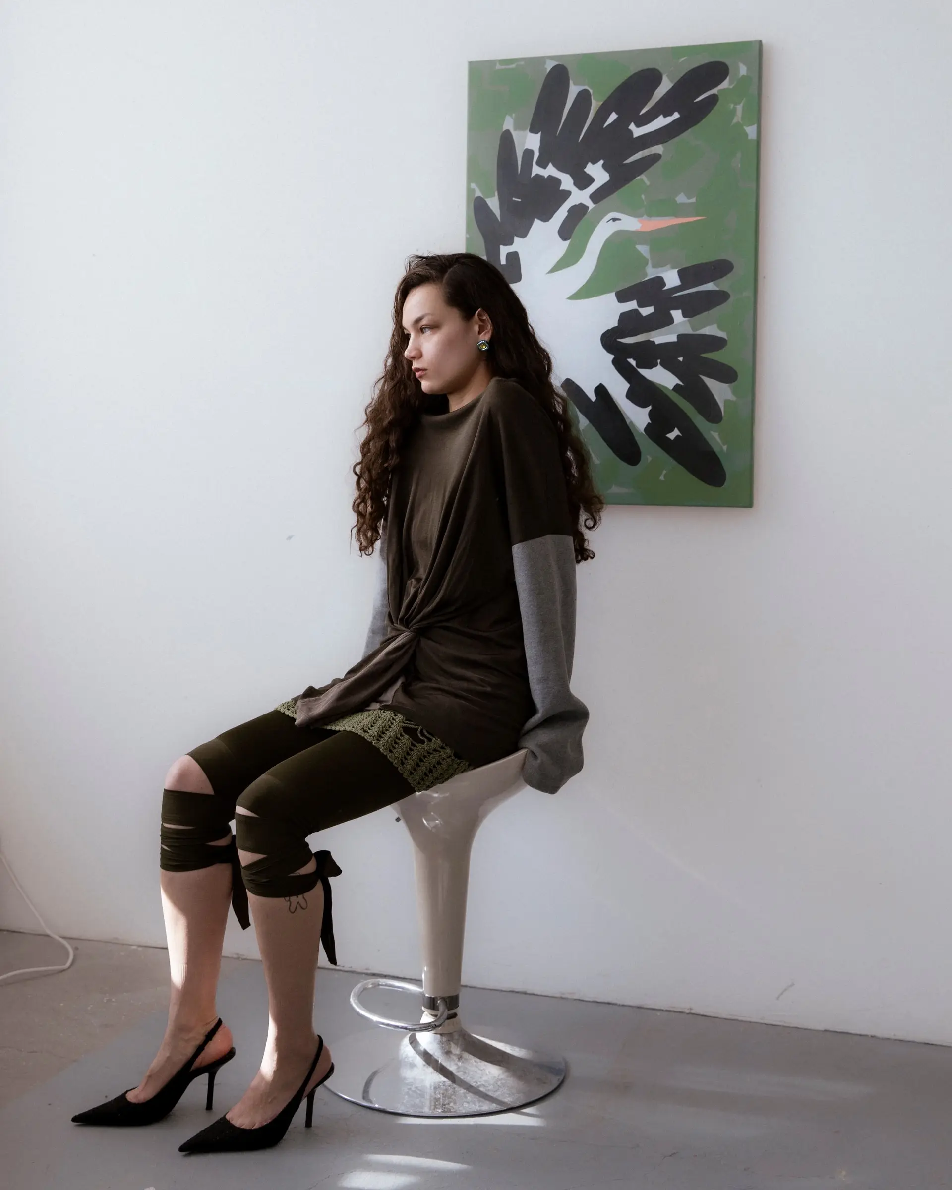 A person with long curly hair in a dark oversized top sitting on a white stool beside a painting of a bird.