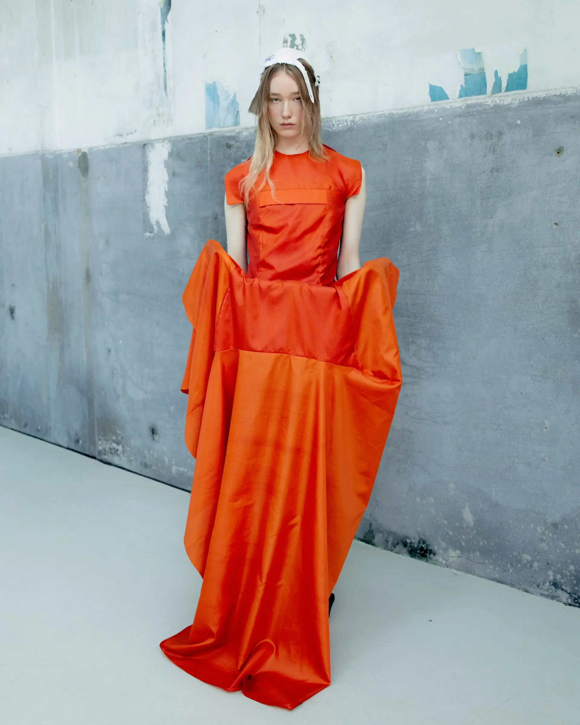 A model in a long draped orange gown in front of a weathered blue-grey concrete wall.