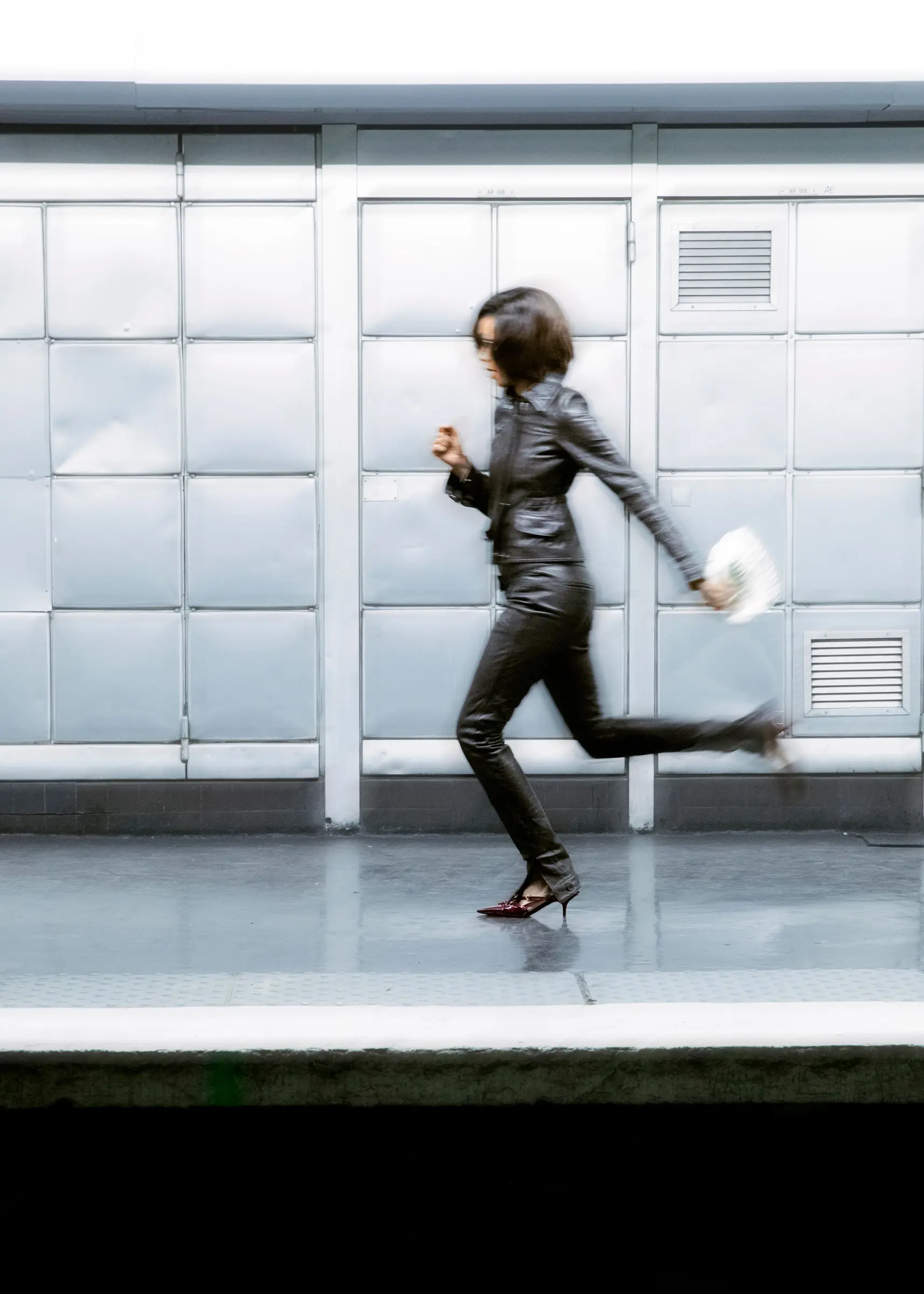 A person in a black leather outfit running past a wall of metal panels, a small white bag in one hand.