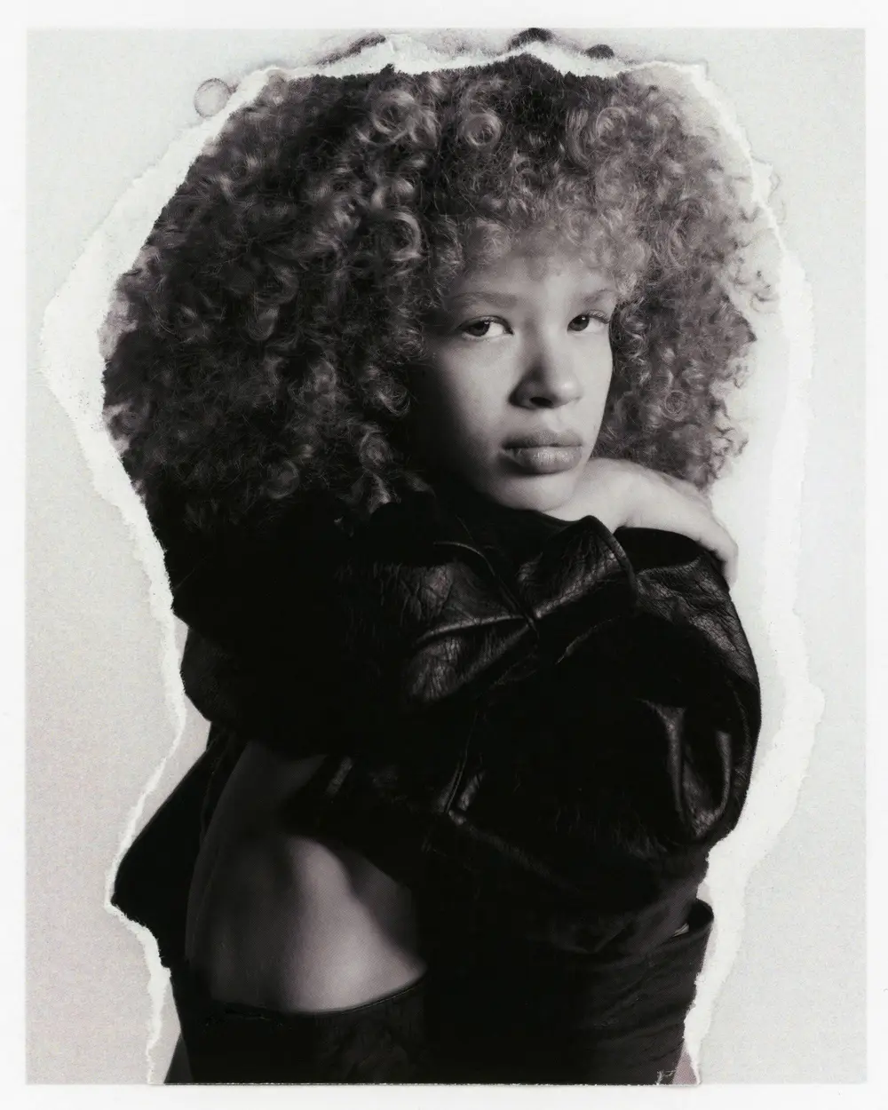 Black-and-white portrait of a person with voluminous curly hair in a leather jacket, on a print with torn edges.