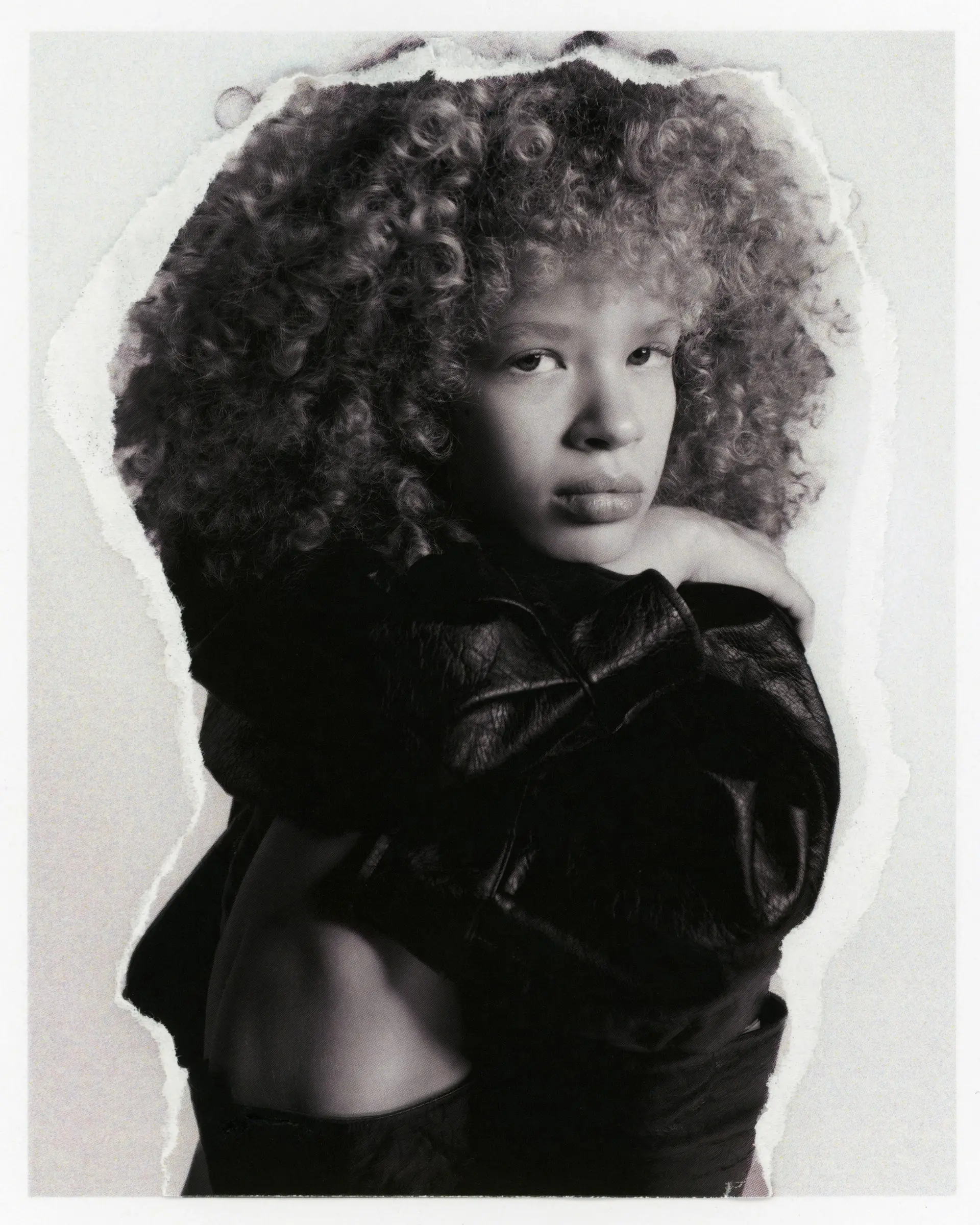 Black-and-white portrait of a person with voluminous curly hair in a leather jacket, on a print with torn edges.