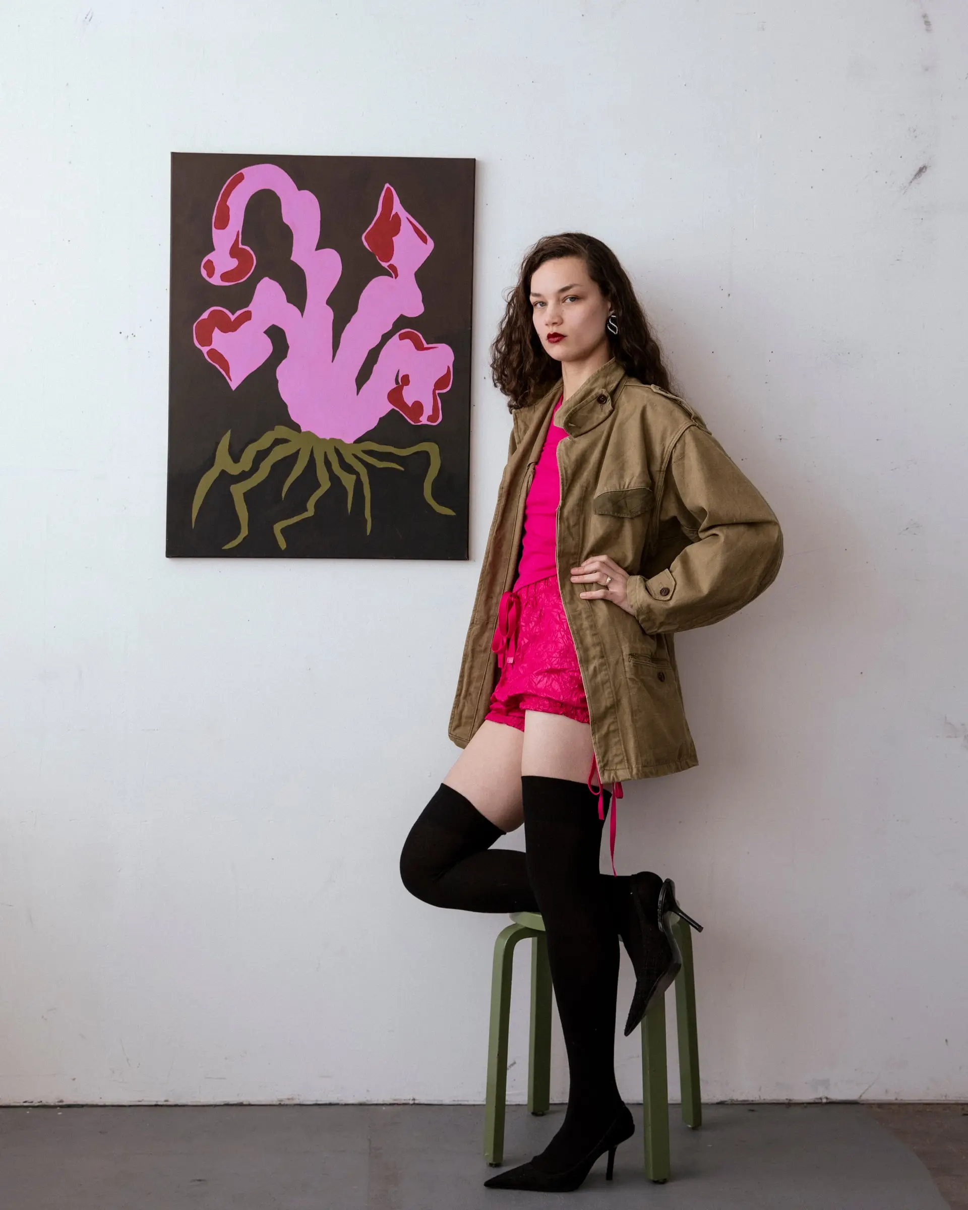 A person in an olive jacket and pink top standing with one foot on a stool beside a painting of a pink flower.