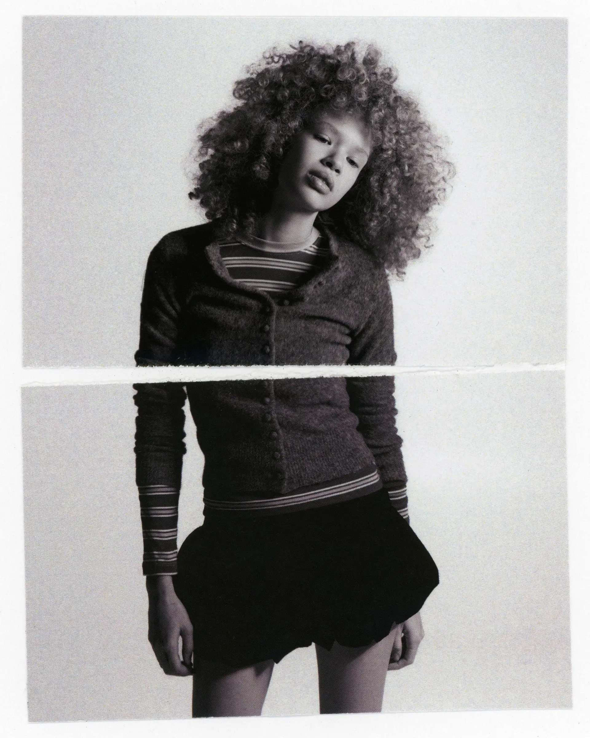 Black-and-white photograph of a person with voluminous curly hair in a cardigan and short skirt, printed on two joined sheets.