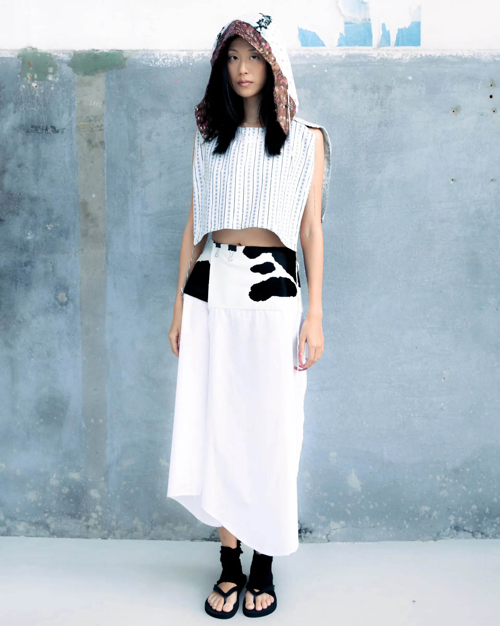 A model in a hood, a knitted cropped top and a long white skirt with black patches against a concrete wall.