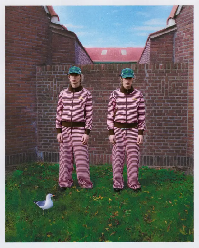 Two men in matching striped tracksuits and caps in front of a brick wall on a lawn.