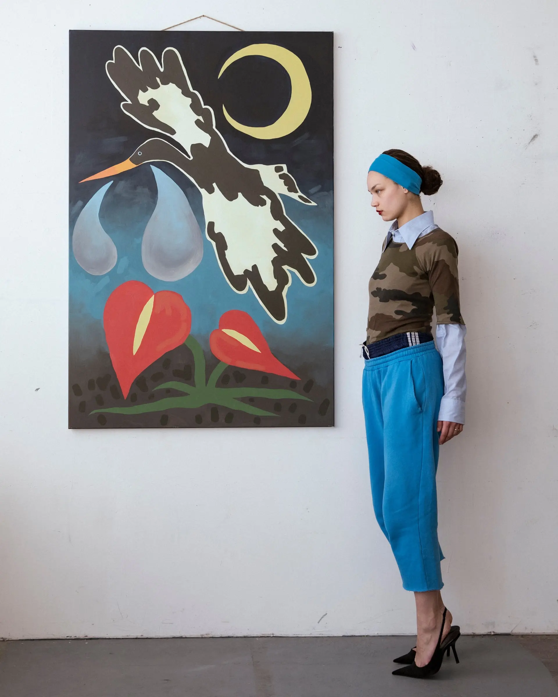 A person in a blue headband, camouflage top and blue trousers beside a large painting of a bird and a crescent moon.
