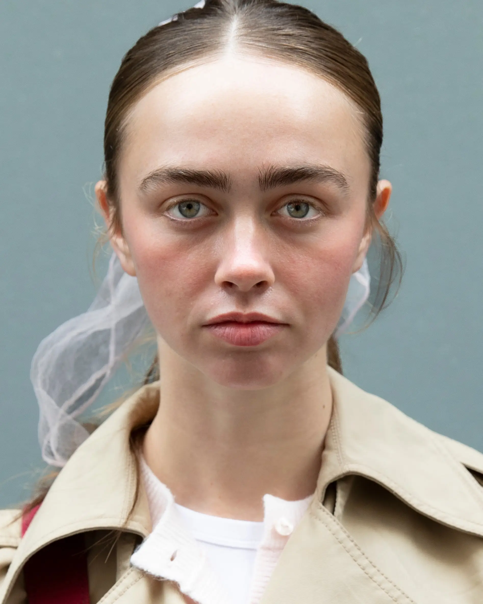 Portrait of a person with hair pulled back and a sheer veil, wearing a beige trench coat.