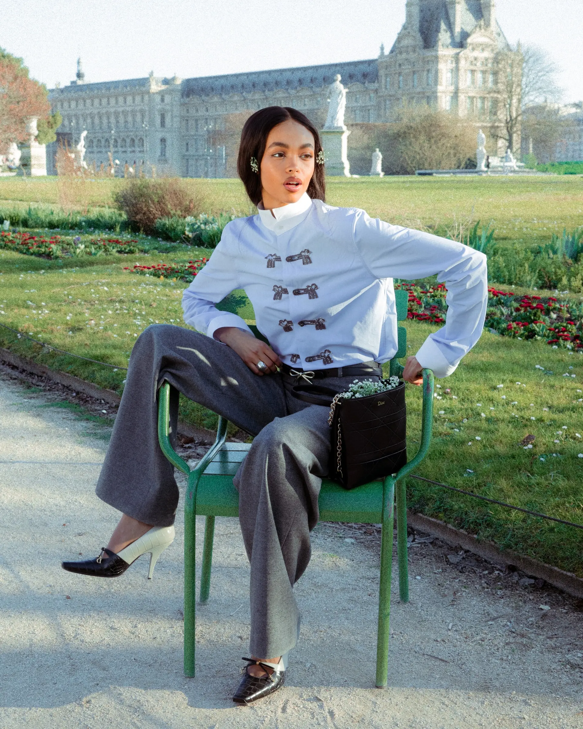 A person in a light blue shirt and grey trousers sitting on a green garden chair on a gravel path.