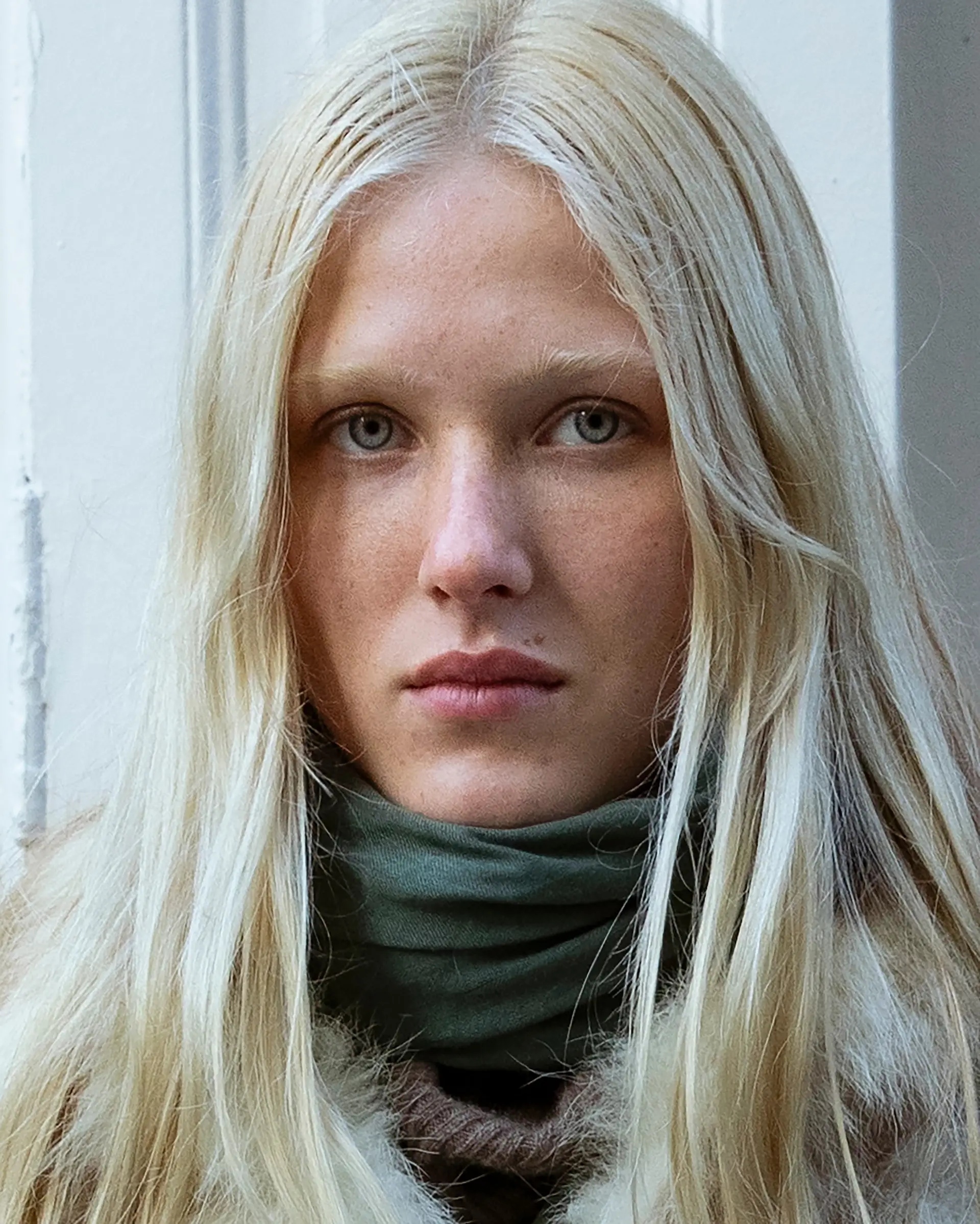 Portrait of a person with long blond hair and a green scarf against a white wall.