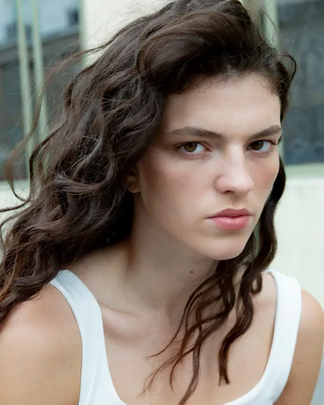Portrait of a person with long dark curly hair in a white tank top, looking into the camera.