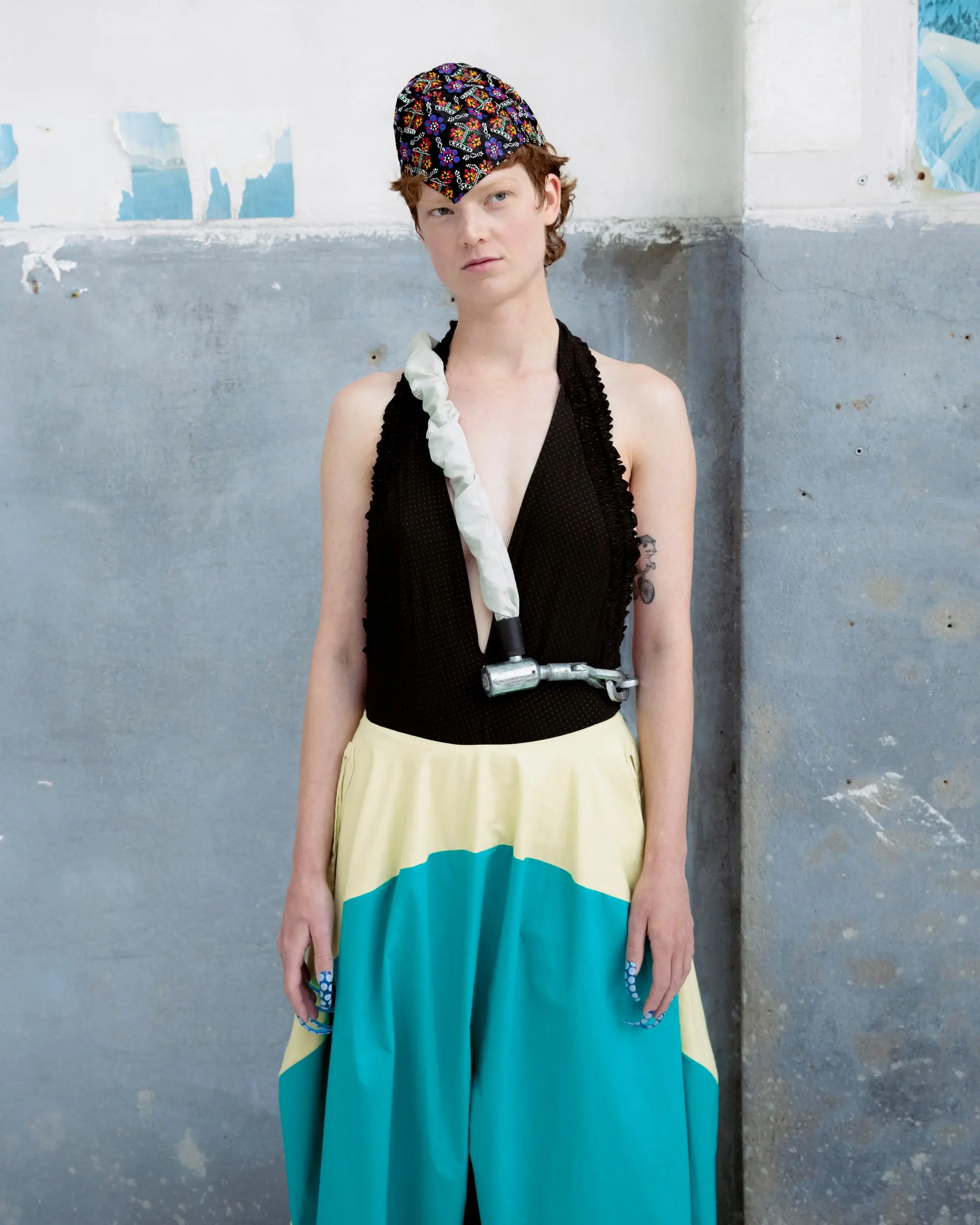 A model in a black halter top, a yellow and turquoise skirt and a patterned cap against a concrete wall.