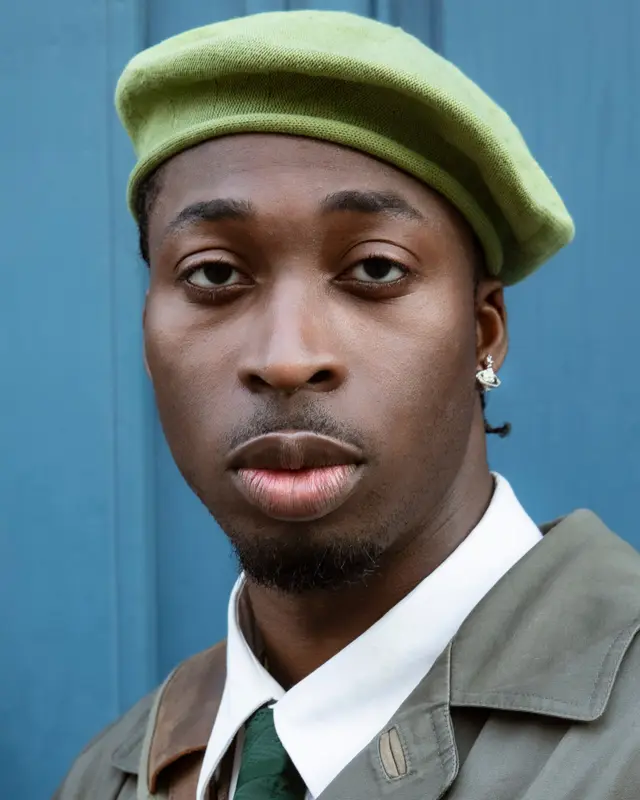 Portrait of a man in a green beret, white shirt and tie, in front of a blue door.
