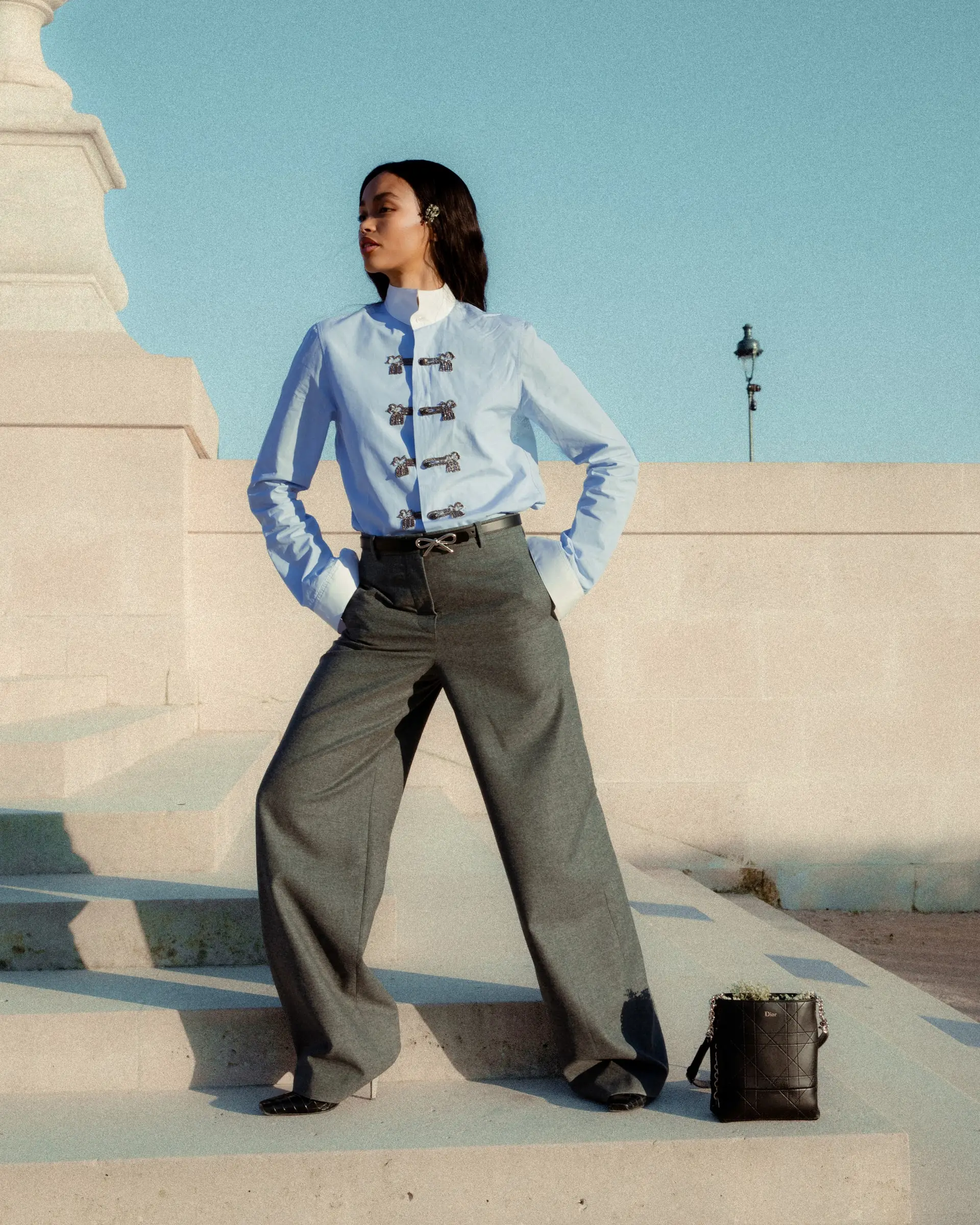 A person in a light blue shirt and wide grey trousers standing on pale stone steps, hands on hips.
