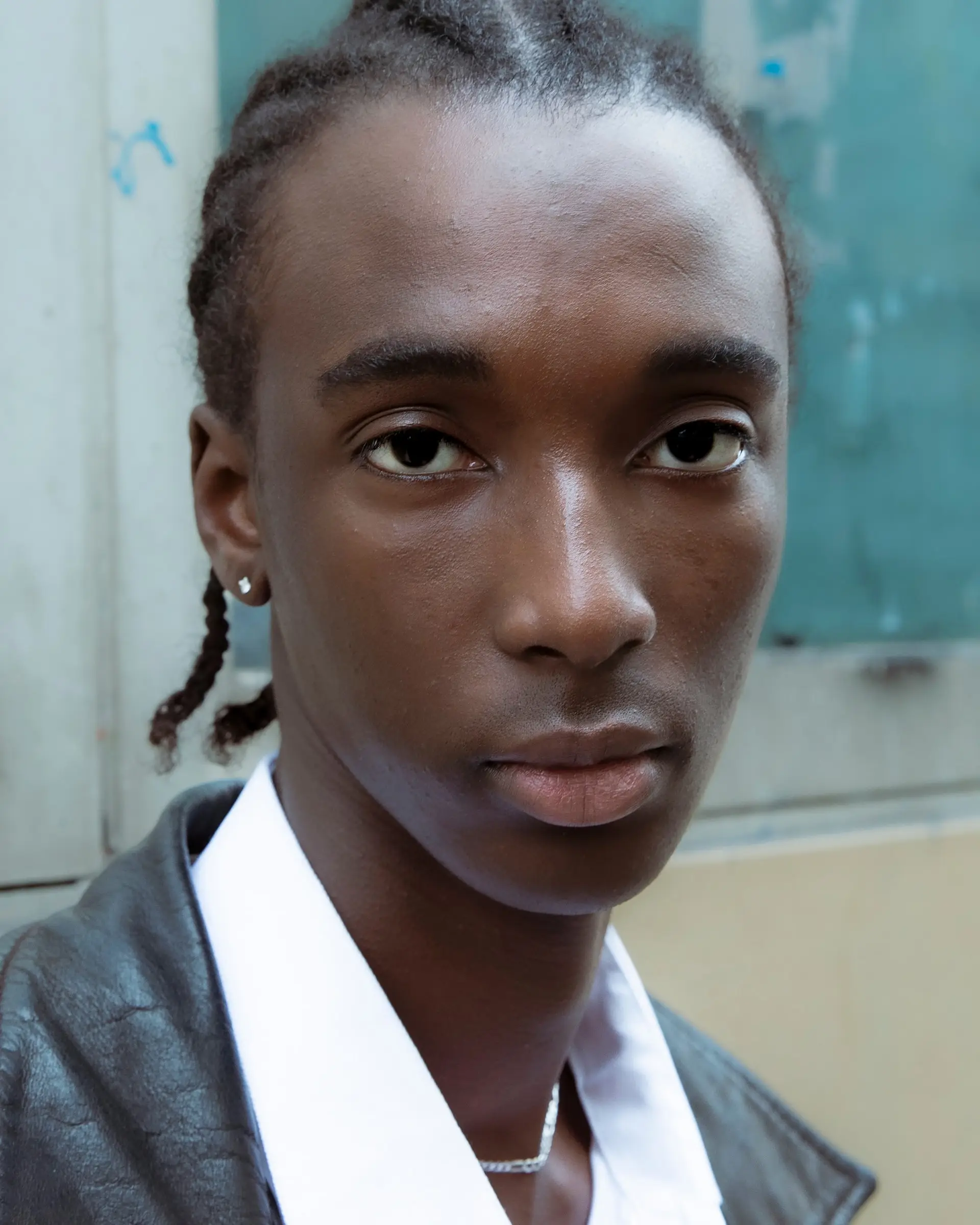 Portrait of a young man with braided hair and a white collar against a pale wall.