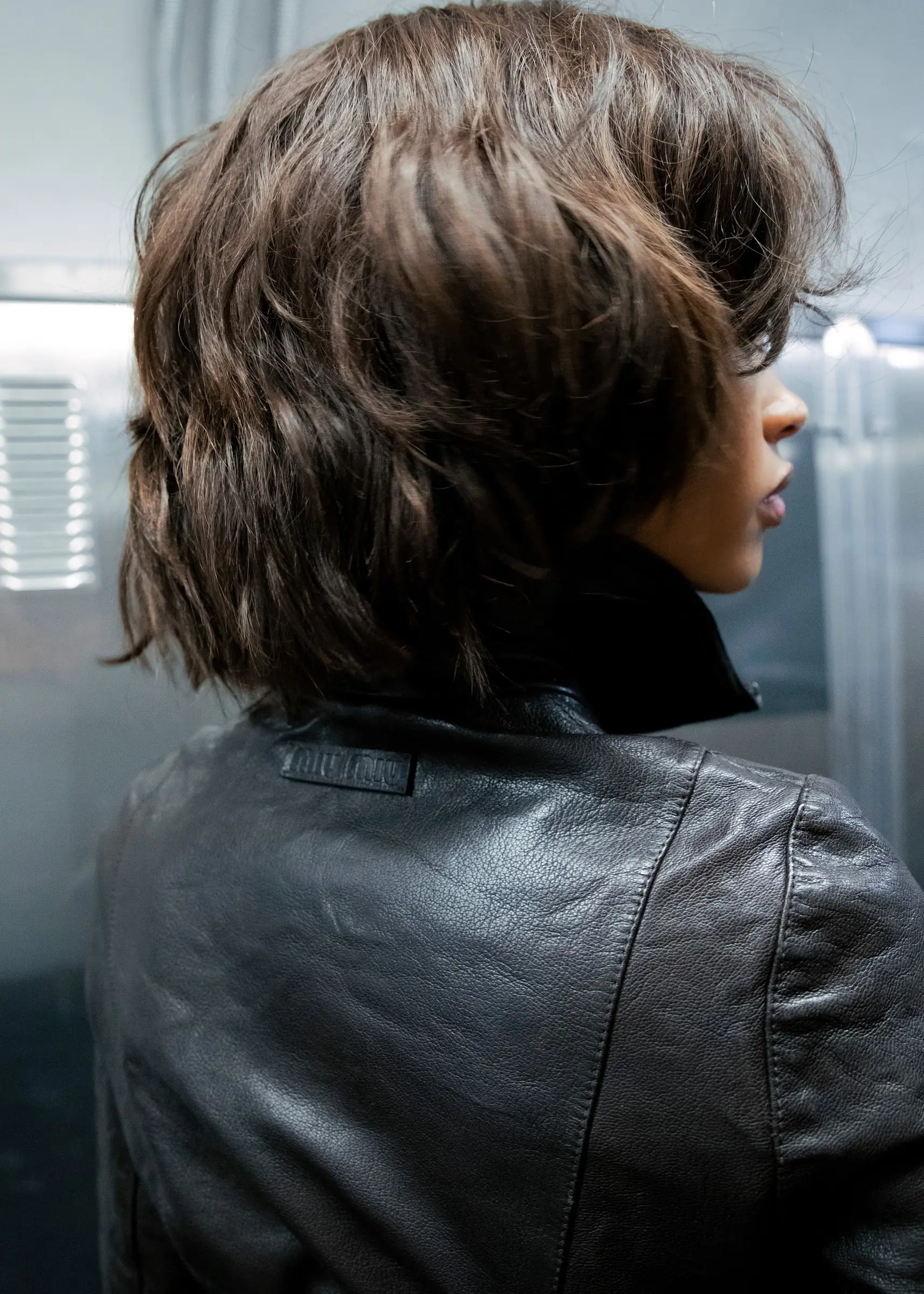 A person with a short dark bob in a black leather jacket, seen from behind.
