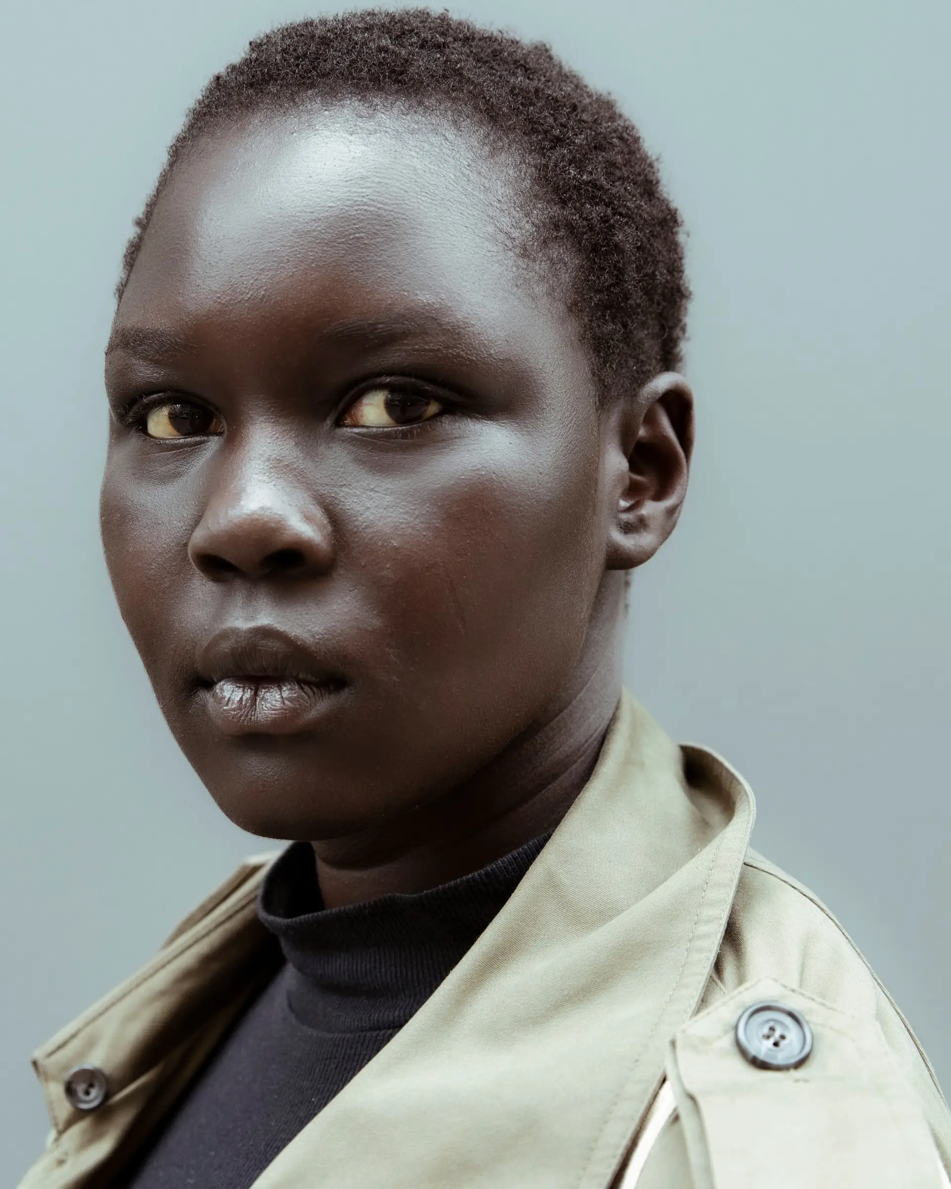 Portrait of a person with short hair in a black top and a beige trench coat, against a grey background.