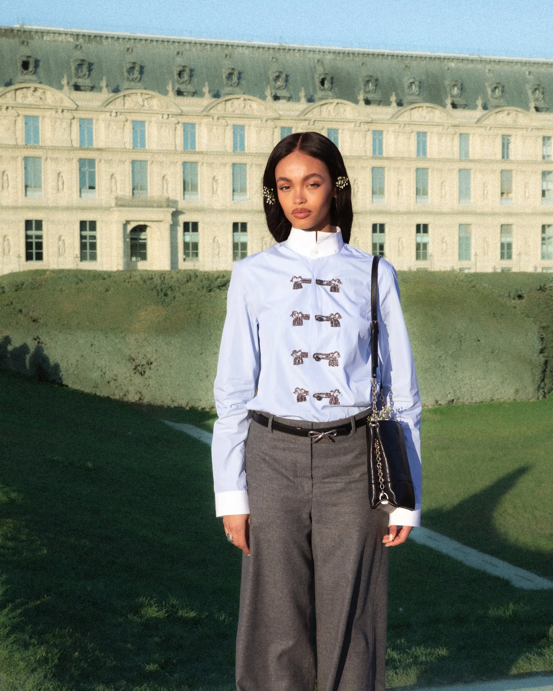A person in a light blue shirt with ornate fastenings and grey trousers in a garden in front of a large classical building.