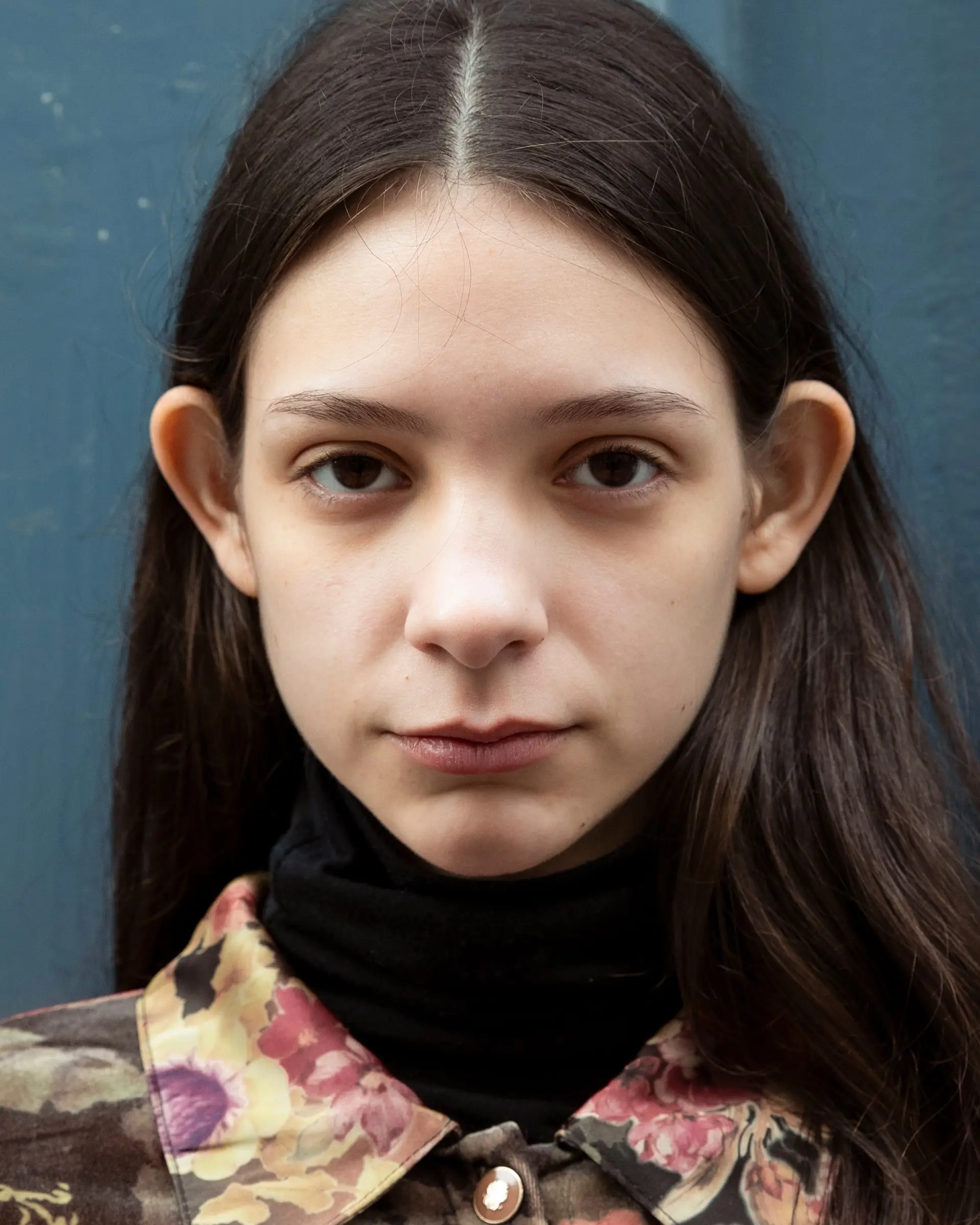 Portrait of a person with long dark hair parted in the middle, in a floral jacket over a black turtleneck.