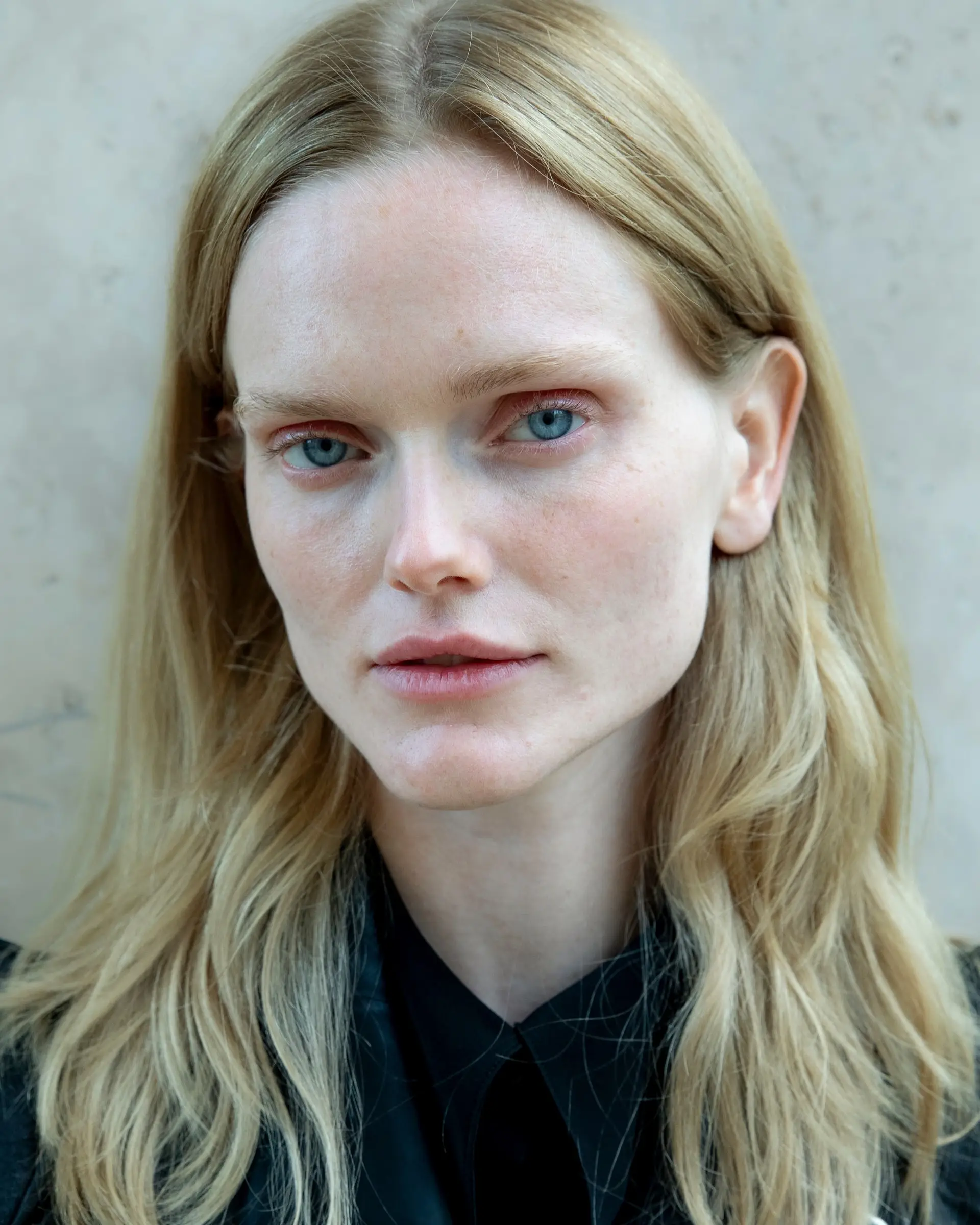 Portrait of a person with long wavy blond hair in a black shirt against a pale wall.