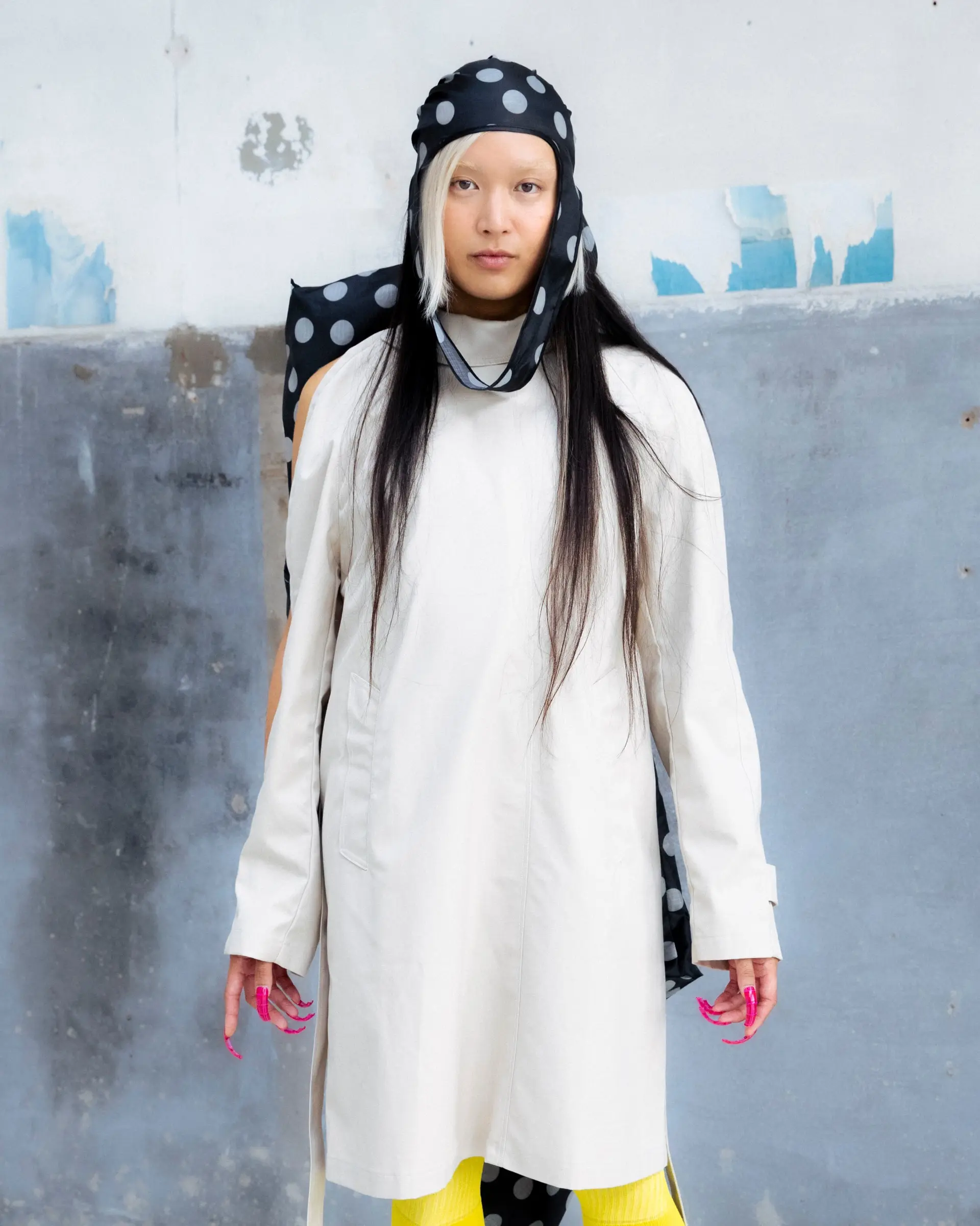 A model in a long white coat and a polka-dot headscarf, with yellow tights, against a concrete wall.